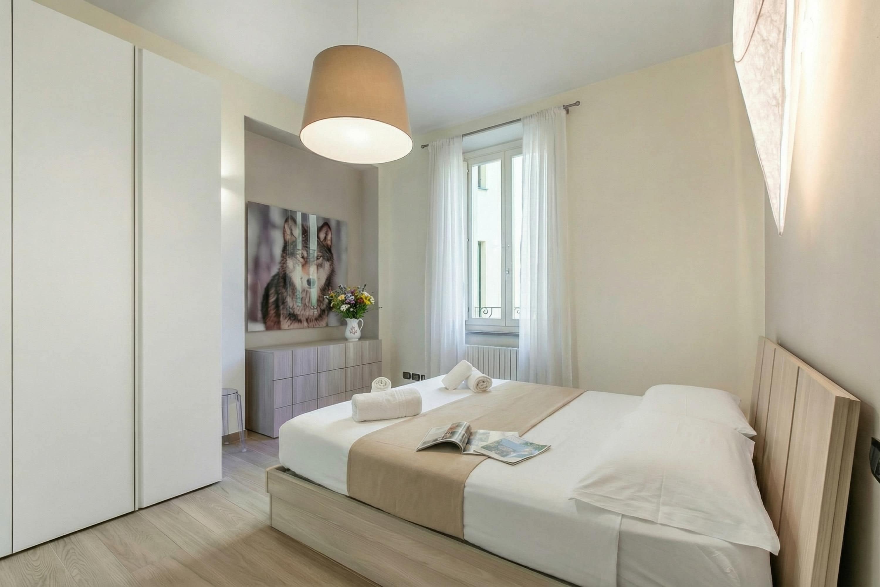 A modern bedroom with a bed, a bedside table, a lamp, a window with curtains, and a painting on the wall.