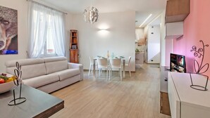 Una sala moderna con sofá, mesa de comedor y sillas, una lámpara colgante y un televisor en la pared.