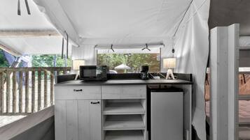 A compact kitchenette with a microwave, coffee maker, and lamp, under a white tented roof with a view of greenery outside.