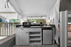 A compact kitchenette with a microwave, coffee maker, and lamp, under a white tented roof with a view of greenery outside.