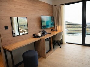 A modern hotel room with a wooden desk, a flat-screen TV, a chair, and a view of a body of water through a large window.