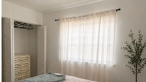 Un dormitorio con cama, cómoda, armario y ventana con cortinas ligeras.