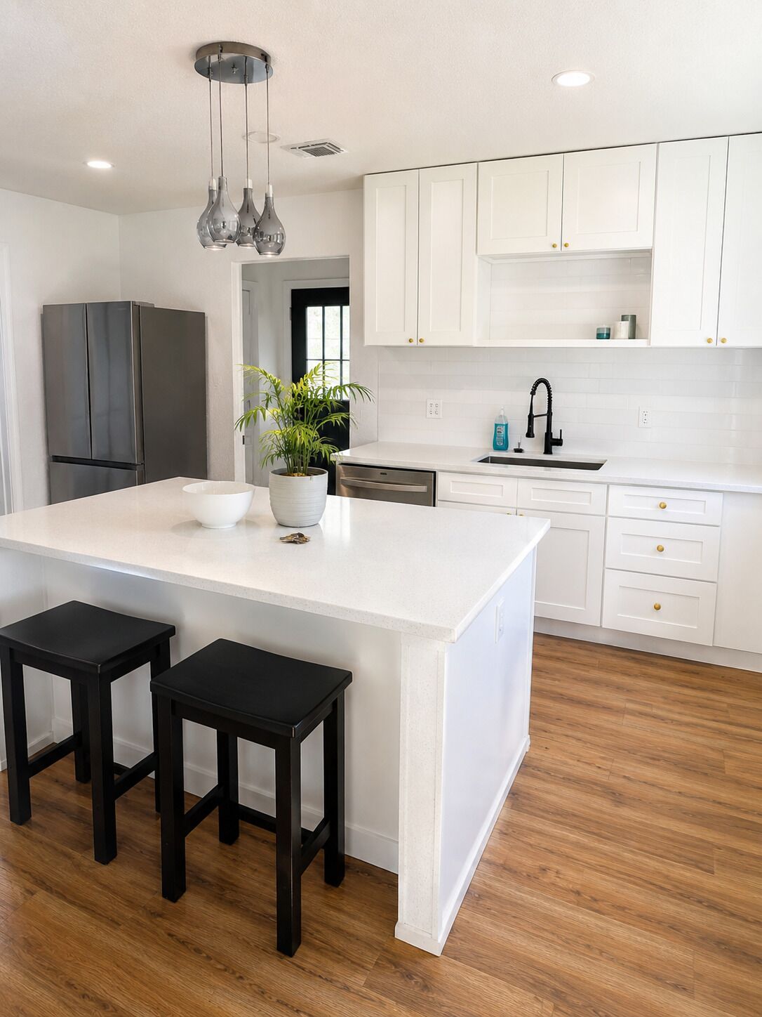 A modern kitchen with white cabinetry, a central island with stools, a stainless steel refrigerator, and a potted plant.