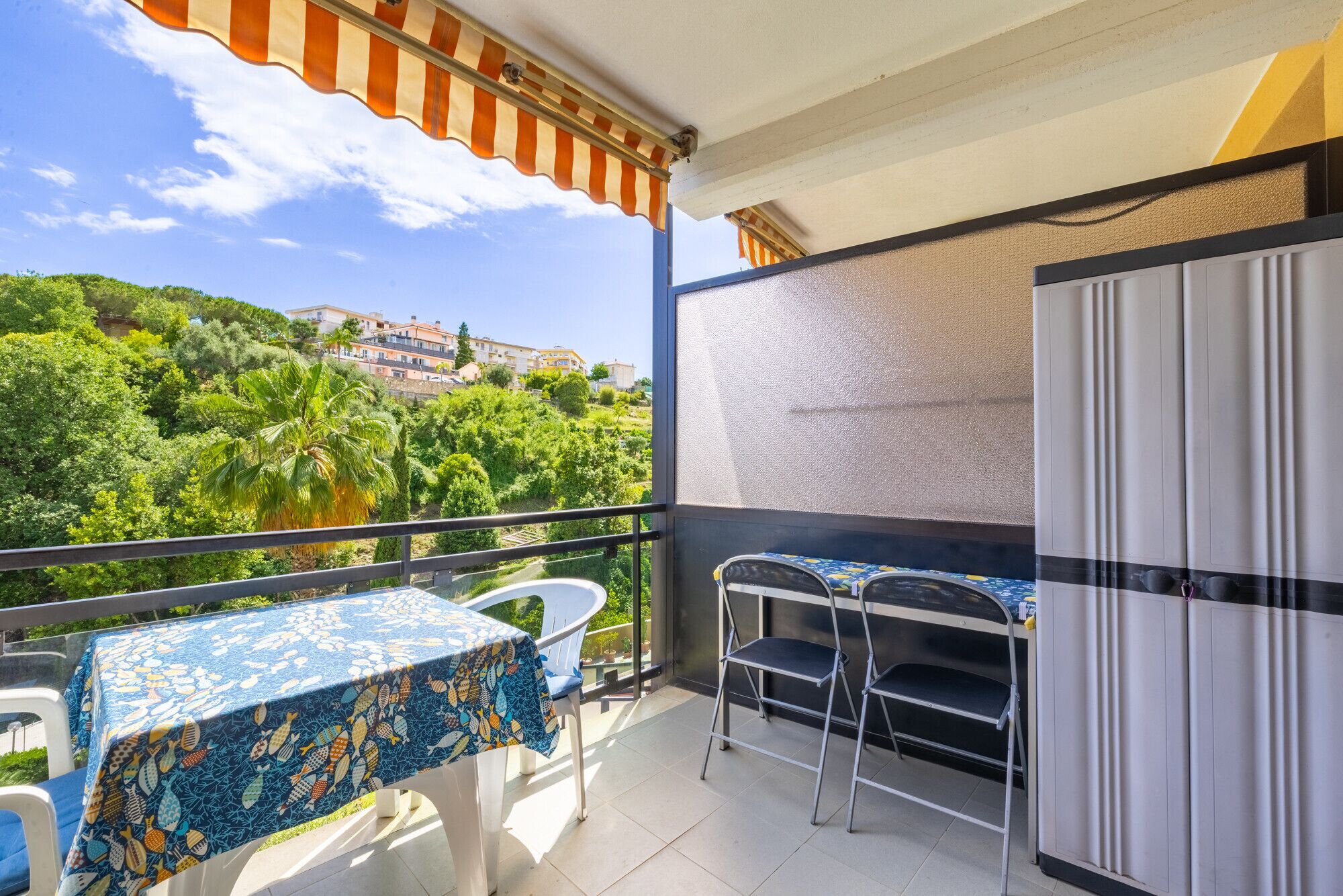A balcony with a table and chairs, a refrigerator, and a view of a green hillside with buildings.