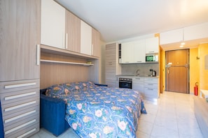 Ein kleines Zimmer mit einem Bett, einer Küchenzeile und einer Tür zu einem weiteren Raum.