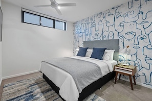 Ein Schlafzimmer mit Bett, Nachttisch, Deckenventilator, Fenster und Wand mit Gesichtsmotiv.