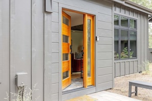 Een gebouw met een grijze gevel, een houten deur met glaspanelen en een raam met uitzicht op groen.