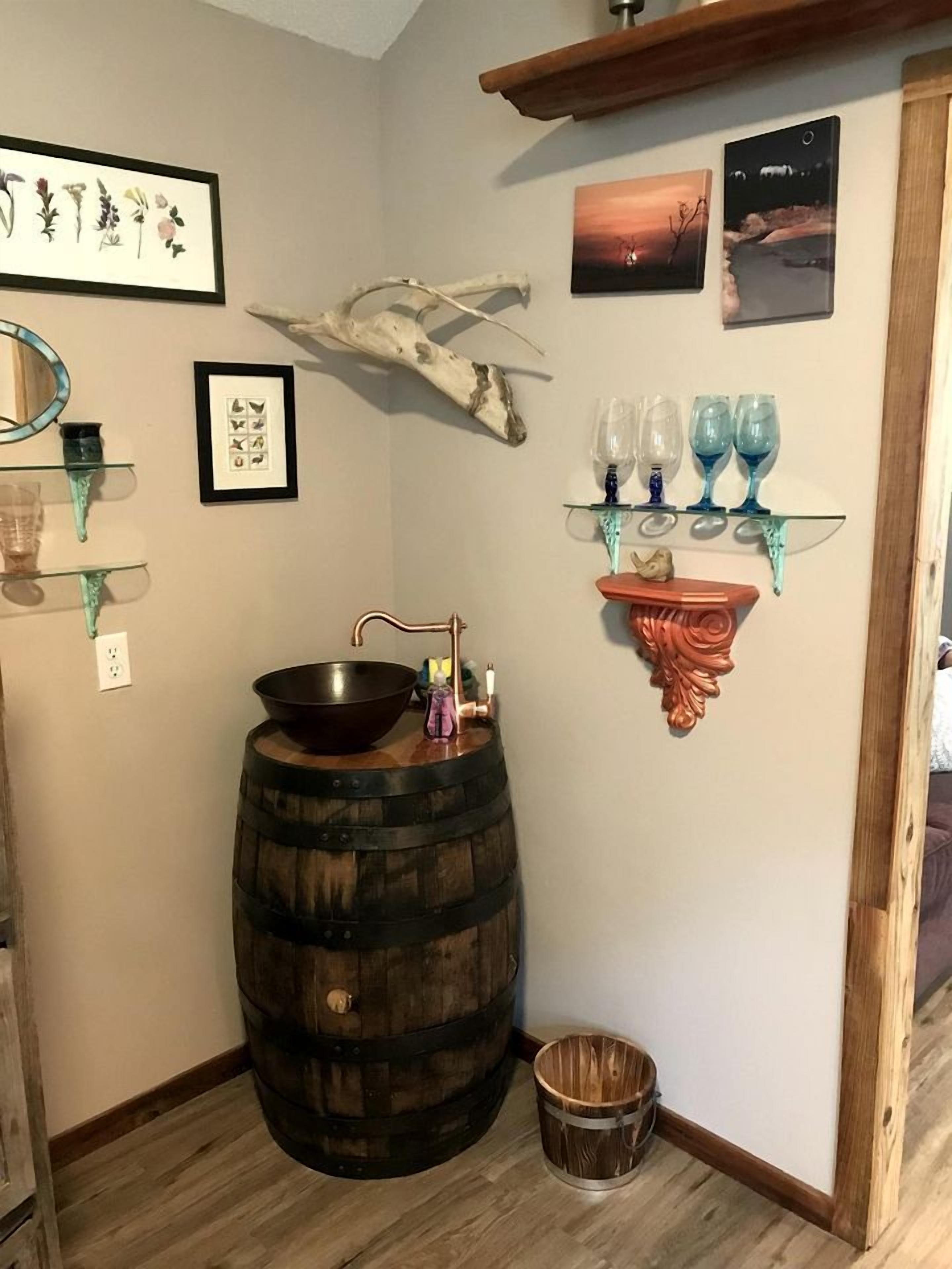 A rustic interior with a wooden barrel sink, a small wooden bucket, a decorative antler, framed botanical prints, and a glass shelf with blue glassware.