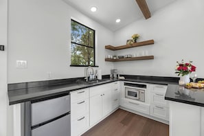 A modern kitchen with white cabinetry, a black countertop, a built-in microwave, and a window with a view of trees.