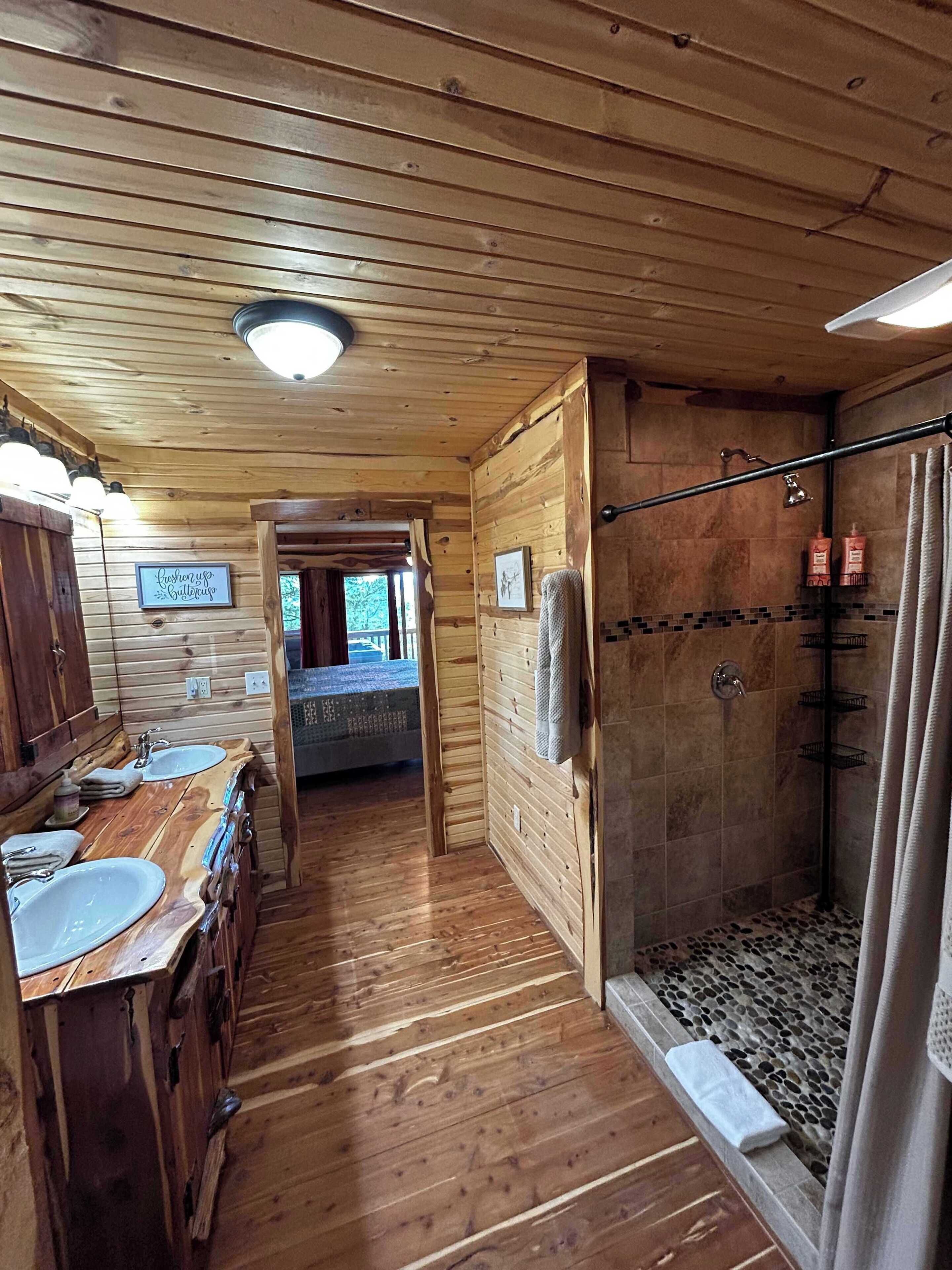 Interior de una cabaña de madera con suelo de madera, un baño con ducha y una cama visible al fondo.