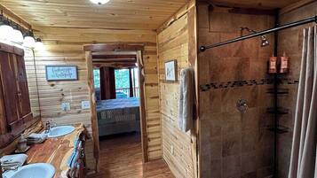 Interior de una cabaña de madera con suelo de madera, un baño con ducha y una cama visible al fondo.