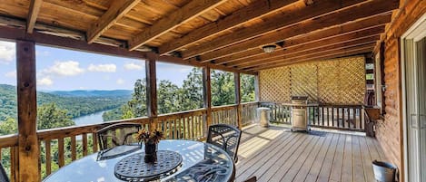 Eine Holzterrasse mit einem Glastisch und Stühlen, mit Blick auf einen Fluss und einen Wald.