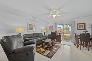 Ein Wohnzimmer mit grauem Ledersofa, einem Glastisch, einem Esstisch mit Stühlen, einem Deckenventilator und Blick ins Freie durch eine Glastür.