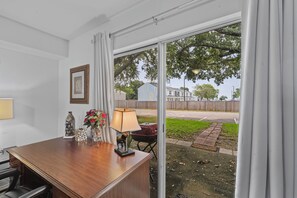 Ein Zimmer mit Holztisch, Lampe, Blumenvase und Blick auf eine Straße und Bäume durch ein großes Fenster.