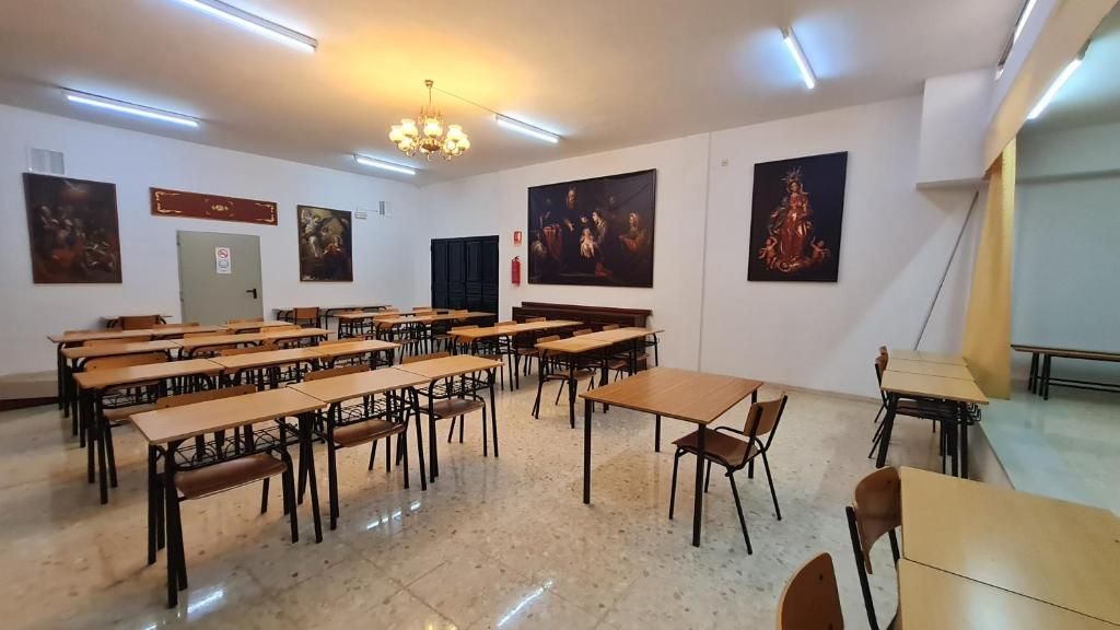 Un aula con escritorios y sillas de madera, una araña y arte religioso en las paredes.