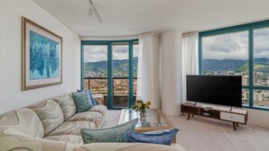 A modern living room with a sofa, a coffee table, a TV, and large windows offering a city view. - 27th-floor Waikiki Suite with Panoramic Mountain/Canal/City Views (Honolulu)