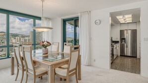 A dining area with a glass table, wooden chairs, a vase with flowers, a view through large windows, and a kitchen with a stainless steel refrigerator. - 27th-floor Waikiki Suite with Panoramic Mountain/Canal/City Views (Honolulu)