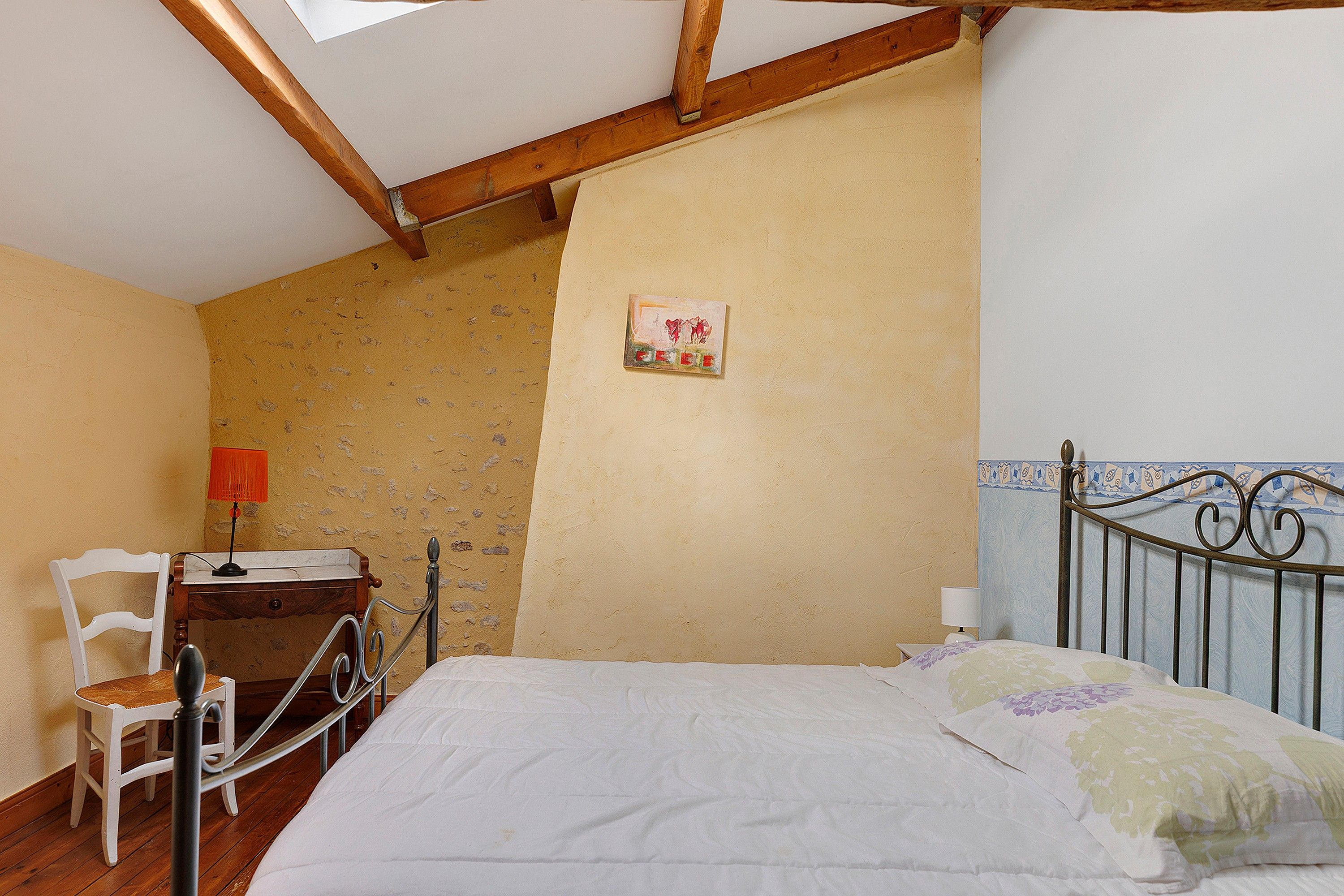 A bedroom with a wooden ceiling, a bed with a floral bedspread, a chair, a desk with a lamp, and a wall with a small painting.