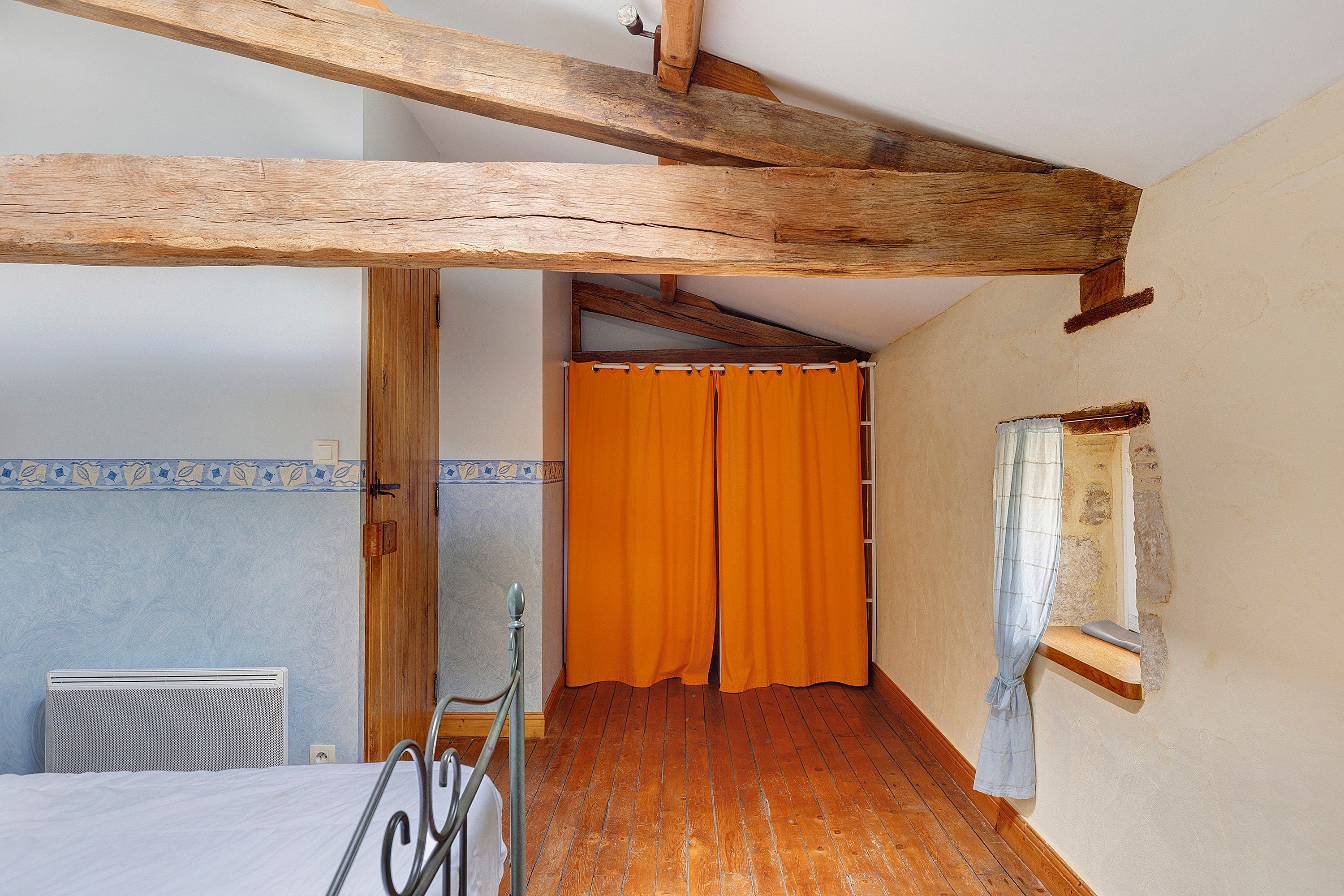 Een kamer met zichtbare houten balken, een bed, een radiator en een badkamer met een oranje douchegordijn.