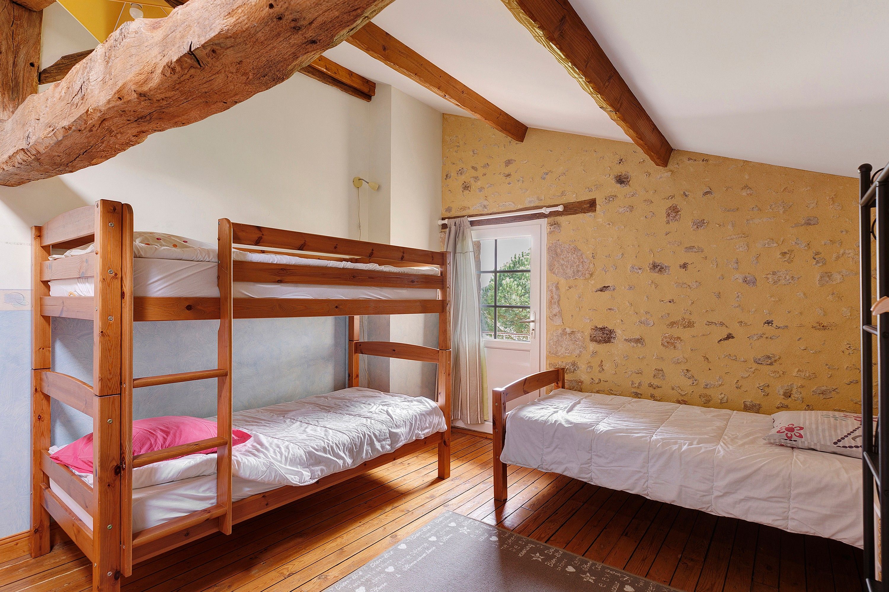 Ein Zimmer mit zwei Etagenbetten und einem Einzelbett, Holzboden, einem Fenster mit Ausblick und einer Steinsäule.