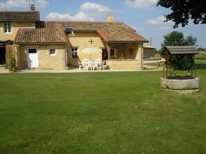 Un bâtiment avec un toit recouvert de tuiles, une croix sur le mur, des places assises à l’extérieur et un petit jardin.