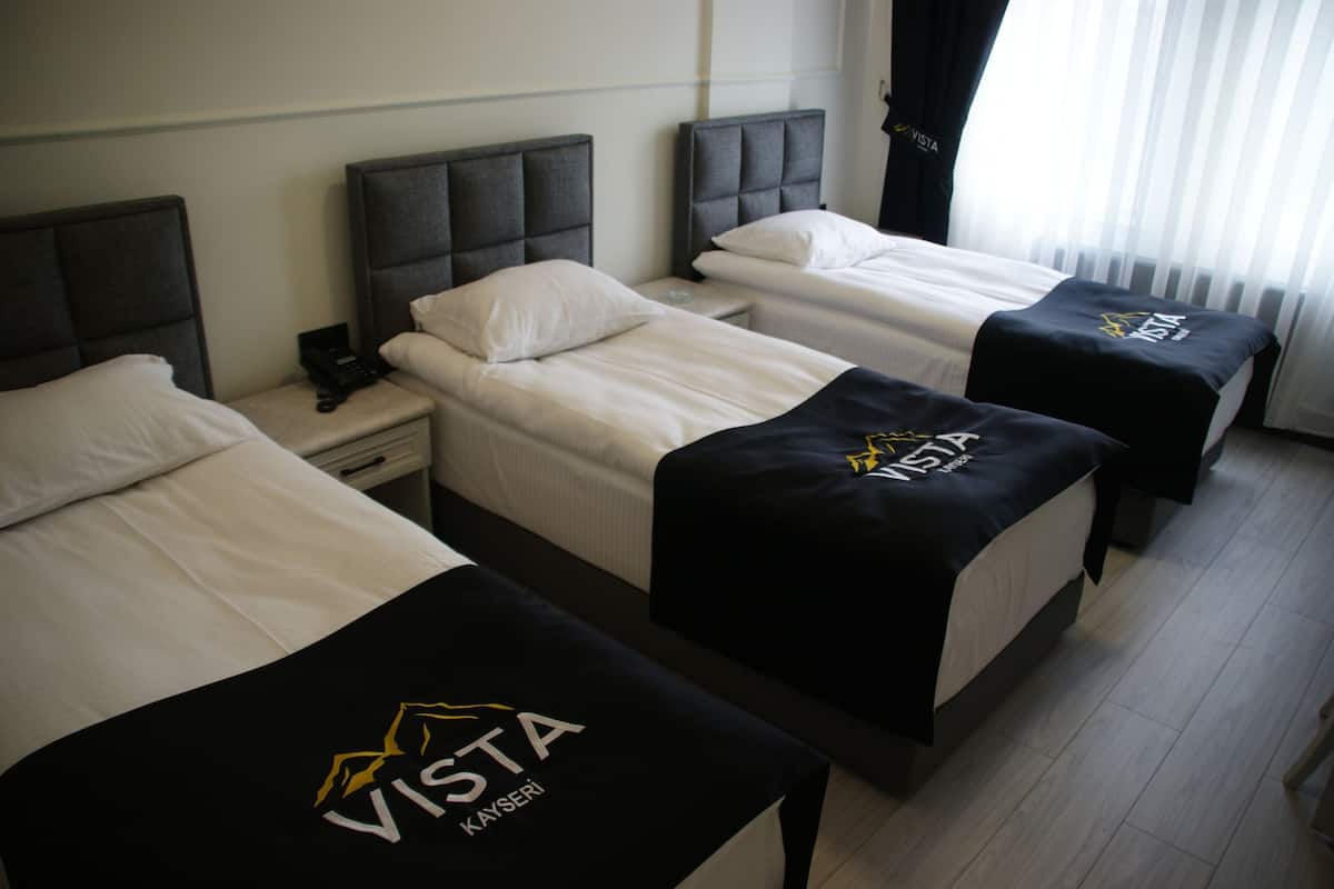 A hotel room with three beds, each with a black and white bedspread featuring the 'VISTA KAYSERİ' logo.