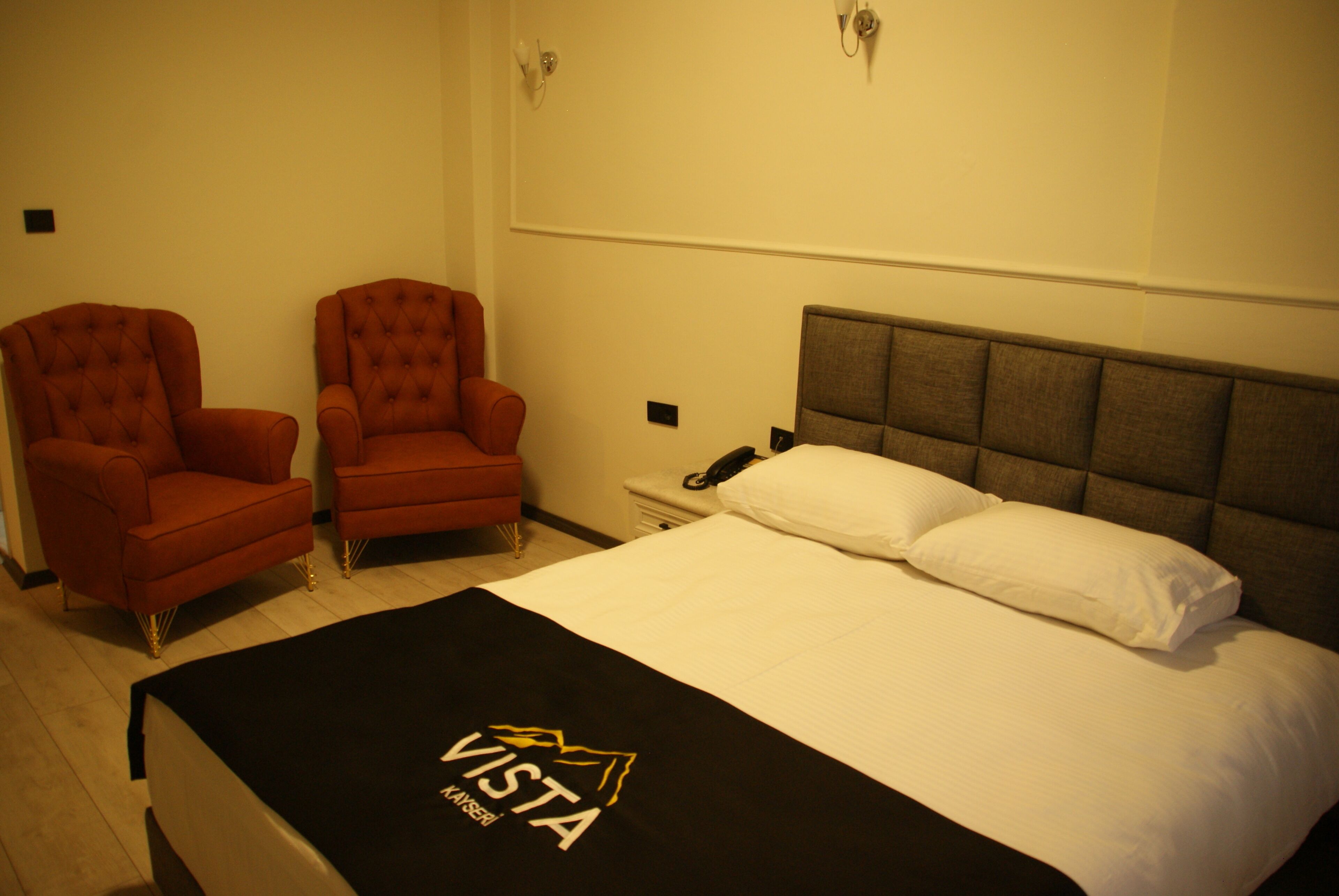 Habitación de hotel con cama, dos sillones, una mesita de noche con teléfono, y una lámpara fijada a la pared.