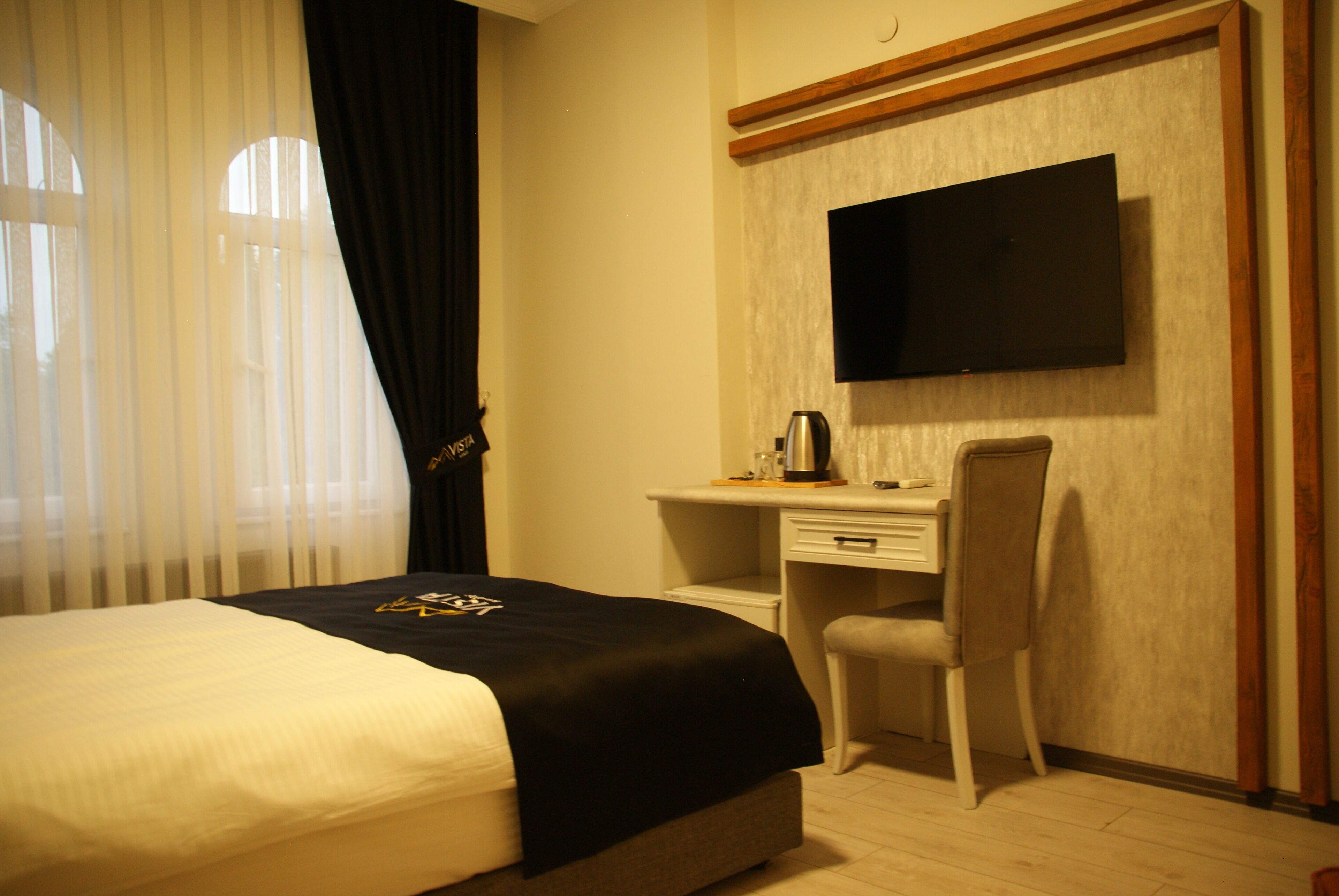 Habitación de hotel con cama, escritorio, silla, televisor montado en la pared y ventana con cortinas.