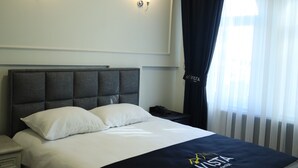 Habitación de hotel con cama, mesita de noche, aire acondicionado de pared y ventana con cortinas de gasa.
