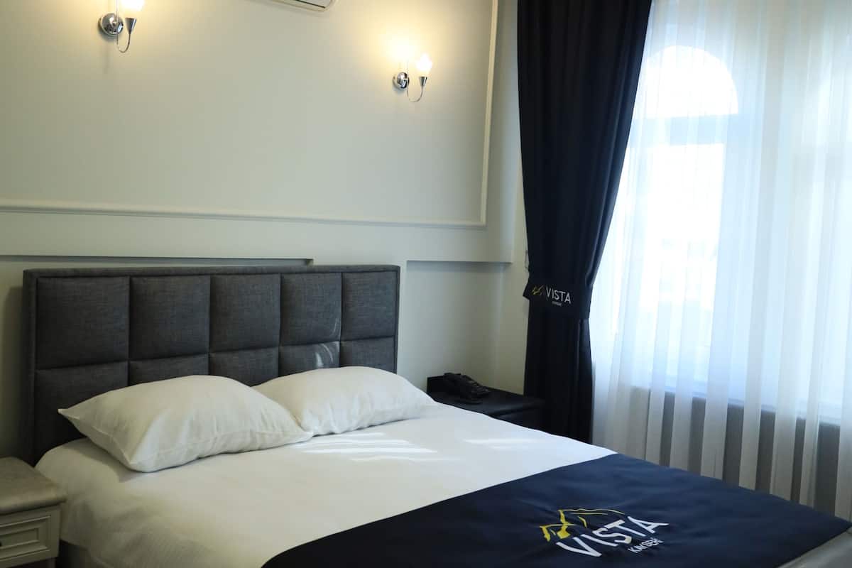 A hotel room with a bed, a nightstand, a wall-mounted air conditioner, and a window with sheer curtains.