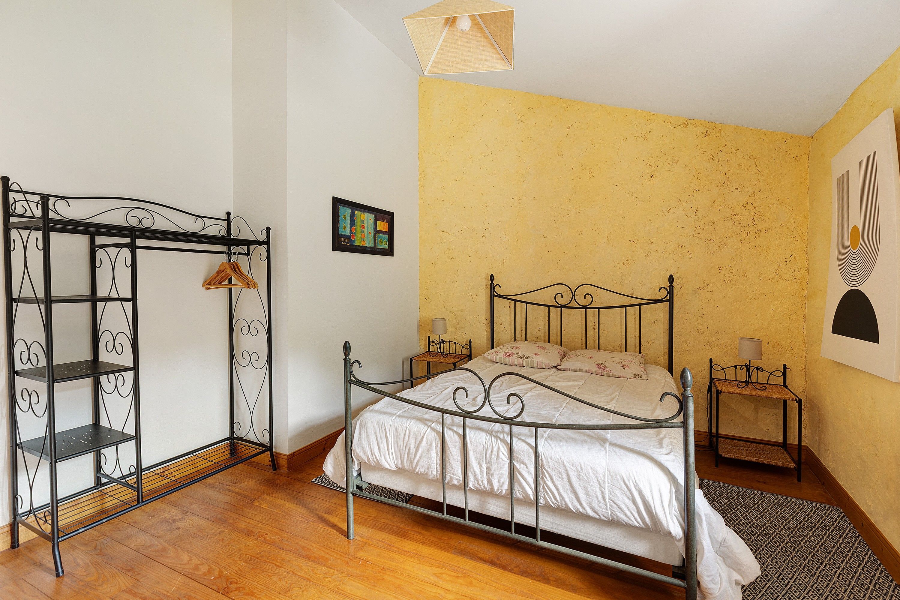 Un dormitorio con estructura de cama metálica, piso de madera, pared amarilla y una mesita de noche con lámpara.