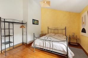 A bedroom with a metal bed frame, a wooden floor, a yellow wall, and a small bedside table with a lamp.