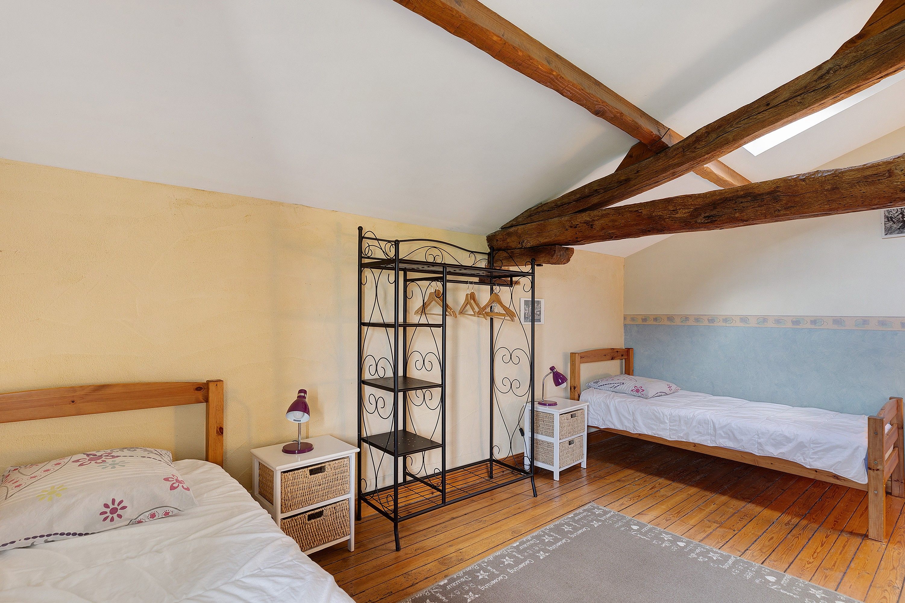 A room with two wooden beds, a metal shelf unit, a small table with a lamp, and a patterned rug.