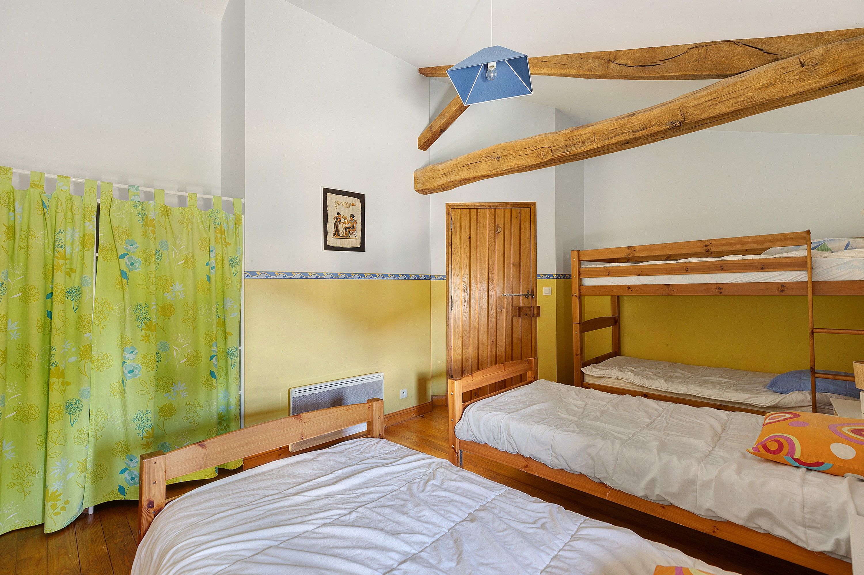 Een kamer met twee stapelbedden, een houten balk in het plafond, een radiator en een groen gordijn met bloemenprint.