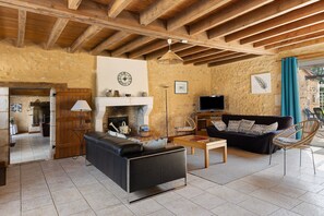 Ein rustikales Wohnzimmer mit freiliegenden Holzbalken, einem Kamin, einer Couch, einem Fernseher und einem Korbstuhl.