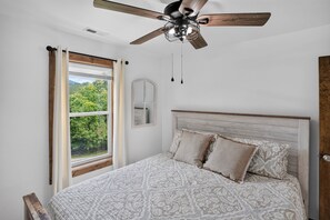 Ein Schlafzimmer mit einem Bett, einem Deckenventilator, einem Fenster mit Vorhängen und Blick auf Grünfläche.