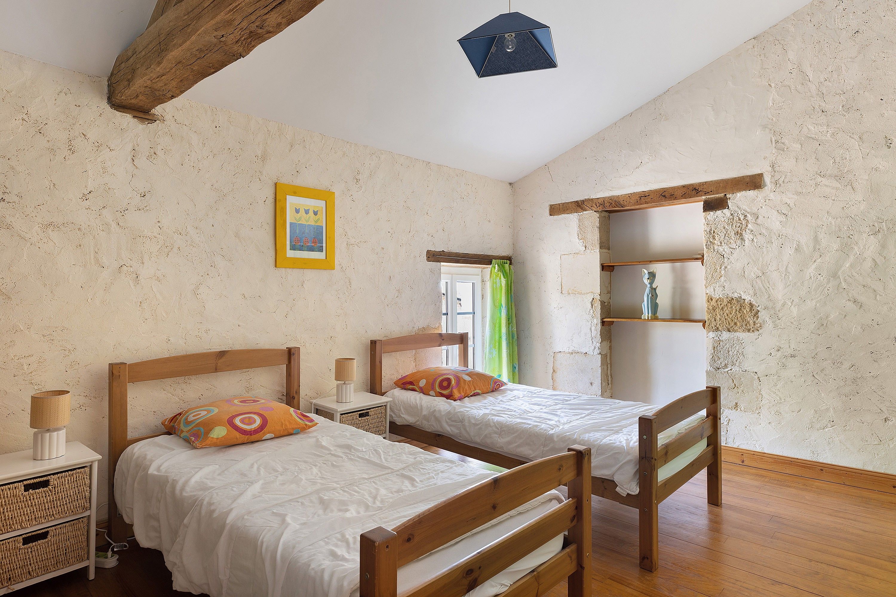 Une chambre à coucher avec deux lits en bois, une table de chevet avec une lampe, un tableau encadré au mur et une étagère avec des objets décoratifs.