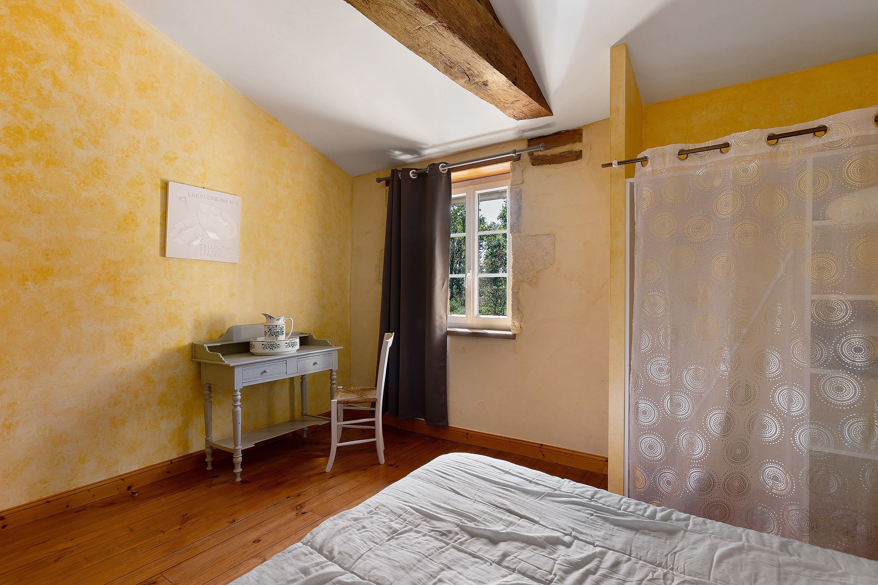 Een slaapkamer met een bed, een bureau, een stoel, een raam met gordijnen en een aan de muur hangend schilderij.