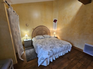 A bedroom with a wooden floor, a bed with floral bedding, a nightstand with a lamp, a wall clock, and a radiator. - Gîte Les Gallinacés: Private Terrace, Shared Garden, and Wi-Fi in Lusignan (Lusignan)
