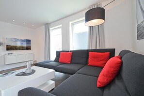 Ein modernes Wohnzimmer mit einem grauen Sofa, roten Kissen, einem weißen Couchtisch und einem großen Fenster mit Vorhängen.