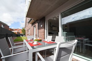 Eine Terrasse mit einem Glastisch und weißen Stühlen, ein Backsteinhaus und Blick auf eine Küche durch eine Glastür.