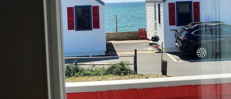 A view of two white buildings with red shutters, a parked car, and a body of water in the background.