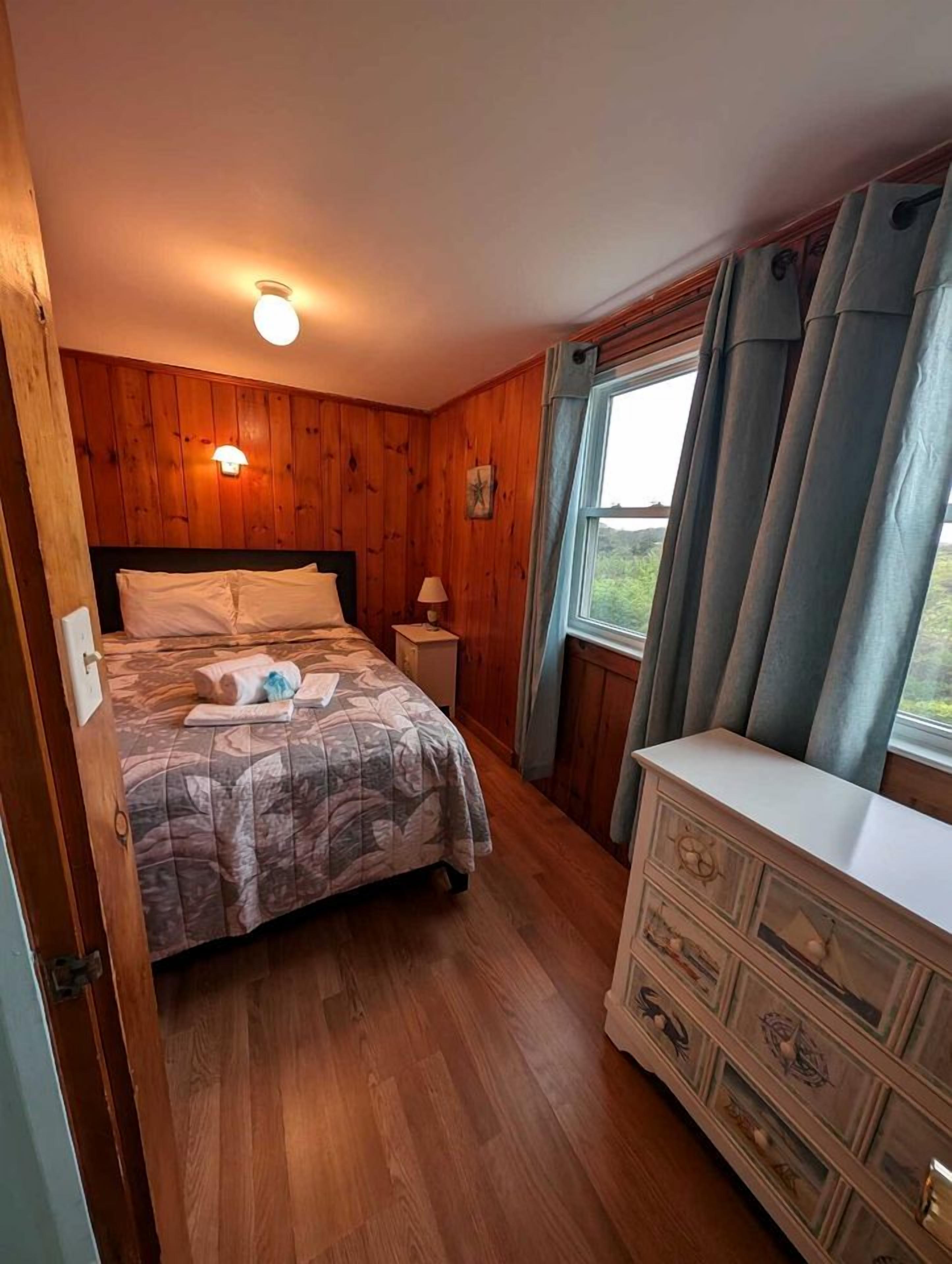 A wooden-paneled room with a bed, dresser, and window with curtains.