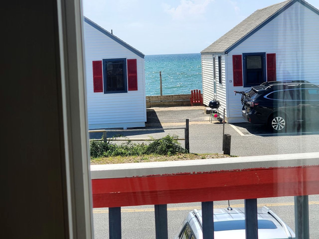 A view of two white buildings with red shutters, a parked car, and a body of water in the background.