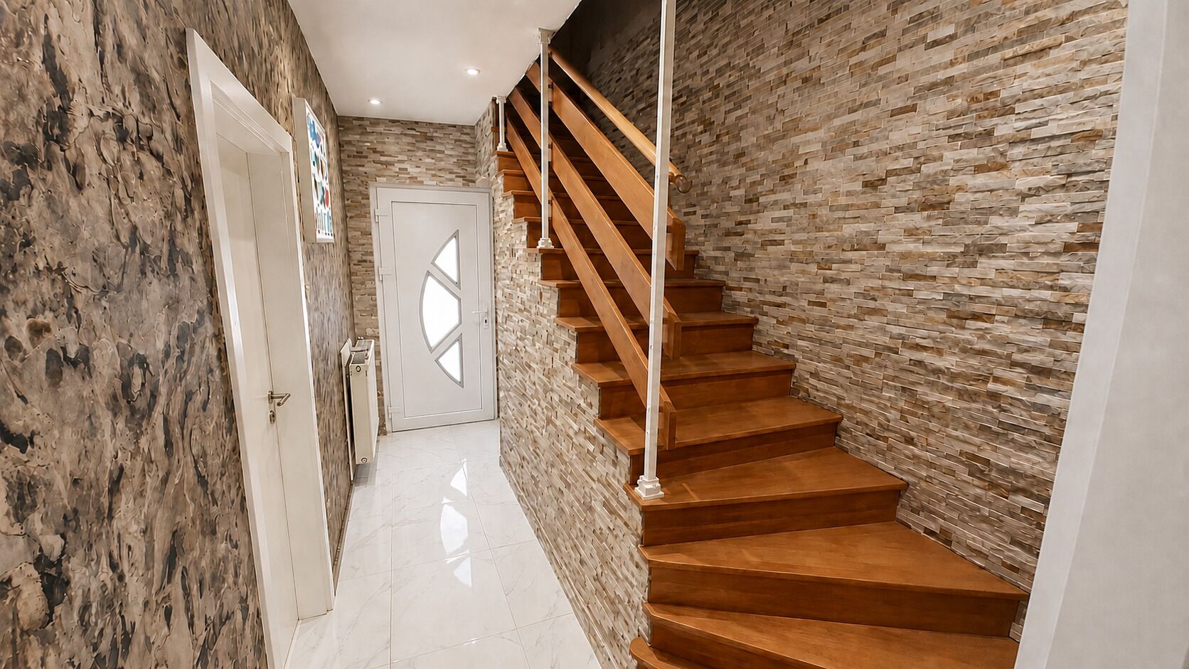Un escalier avec des marches en bois et une rampe blanche, menant à une pièce dont un mur est en pierre et qui possède une porte ornée de motifs géométriques en verre.