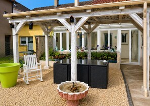 Un patio couvert avec une table et des chaises, un abreuvoir pour oiseaux et un jardin agrémenté de plantes et de fleurs.