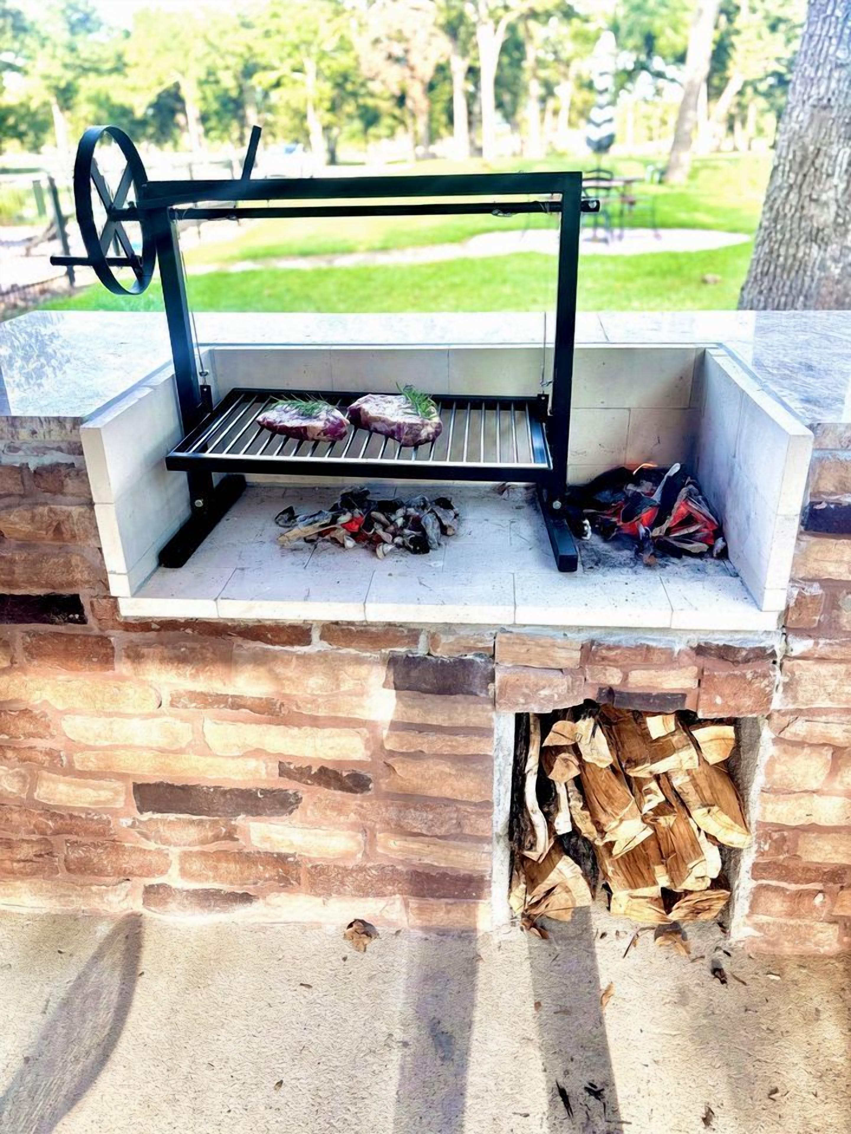 A brick-built outdoor grill with a metal frame, a fire pit below, and a grassy park in the background.