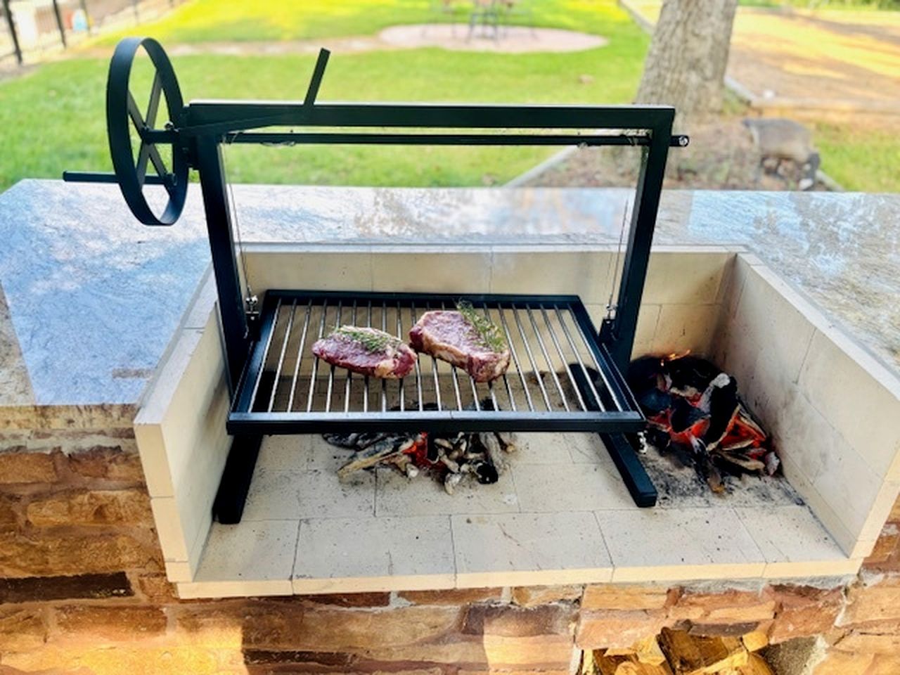 A portable grill with two steaks cooking over an open flame, set against a stone wall.