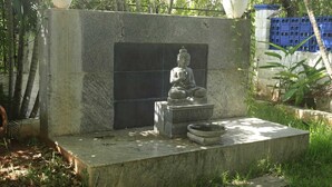 A stone Buddha statue on a pedestal with a water basin in a garden setting.