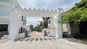 A gated entrance with a white pergola, a paved pathway, and buildings with balconies and windows.